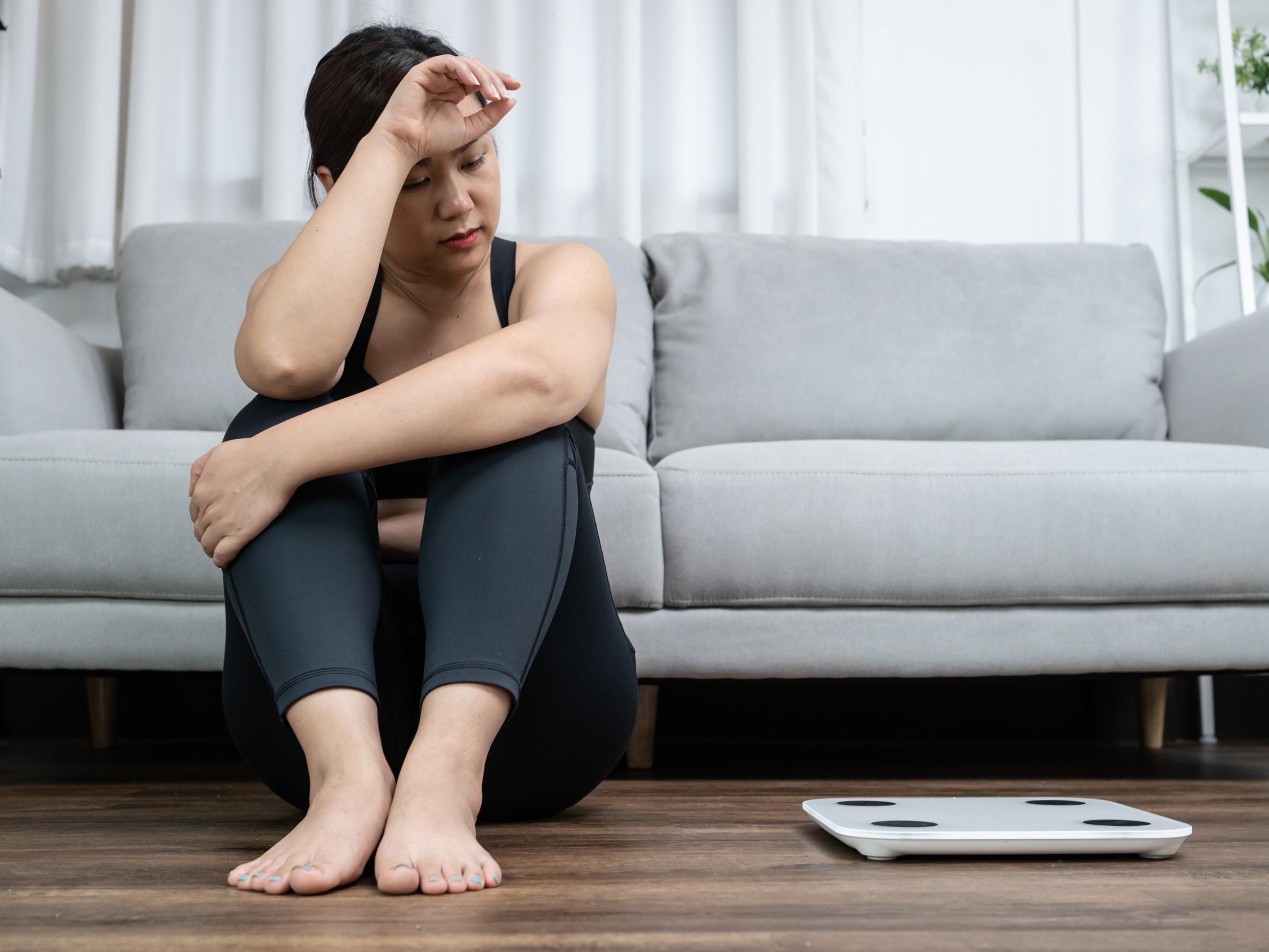 Woman sitting on the floor looking frustrated beside a scale, representing how hormonal imbalance can affect weight loss efforts.